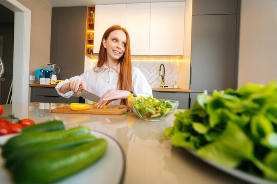 Low-angle view of young redhead woman cutting fresh yellow bell pepper preparing food salad sitting at table in modern kitchen room. Pretty female cooking vegetarian dieting salad full of vitamins.