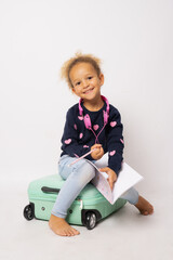 Beautiful little girl sitting on a suitcase and holding a notebook isolated over white background.