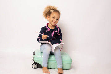 Cute little girl sits on a suitcase and writing in a notebook isolated over white background.