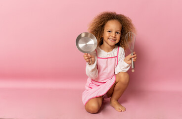 Smiling little girl wearing apron with kitchen utensils isolated over pink background.