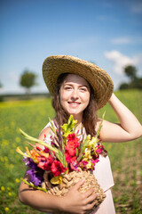 Portrait of smiling white young woman with long hair in light dress in the countryside in straw hat with flowers in her hands