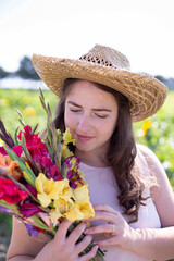 Portrait of white young woman with long hair in light dress in straw hat with bright bouquet of flowers in the countryside