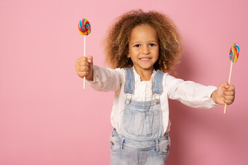 Funny child with candy lollipop, happy little girl eating sugar lollipop isolated over pink background.