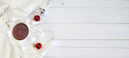 Cup of tea or coffee, knitted white blanket, garland and Christmas balls on wooden background. banner