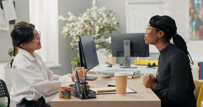 Women sit at desk across from each other job interview common language topic for gossip dark-skinned boss laughs along with candidate for position in corporate office shake hands as sign of agreement