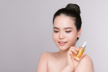 Beauty portrait of an attractive young woman standing isolated over gray background, showing bottle with facial oil
