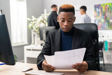 A young boy with unusual hair sits on a chair at a desk in a company, in his hands he holds reports, documents, reads carefully, analyzes the notes, focused, attentive, serious employee in a jacket