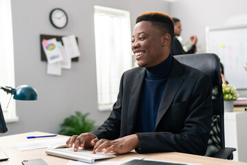 A smiling dark-skinned boy spends morning at work in front of a computer screen, tapping fingers on the keyboard, filling out official and company applications, replying to emails, writing reports