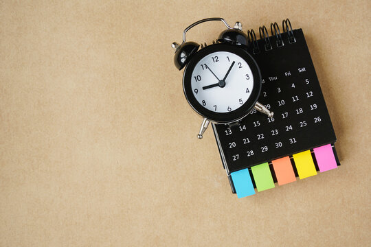black analog alarm clock on opened calendar with multi color paper tag on grunge brown paper background