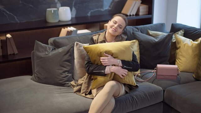 Portrait of smiling happy Caucasian slim beautiful woman admiring and hugging decorative mustard cushion sitting on couch in furniture showroom. Attractive female buyer making choice in shop