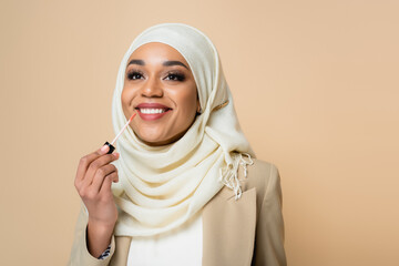 happy muslim woman in hijab applying lip gloss isolated on beige