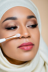 close up of young muslim woman in hijab holding cosmetic brush near face isolated on beige