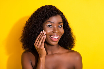 Photo of young happy excited smiling afro girl with naked shoulders apply cream on cheek isolated on yellow color background