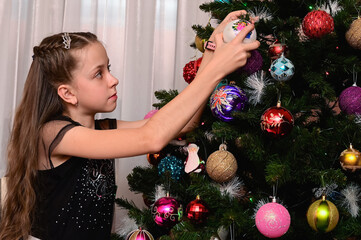 beautiful young girl with long blonde hair holds a Christmas tree toy in her hand and decorates the...