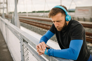 Young blonde runner man checking his smart watch after a running session on the bridge listening to music on his blue headphones