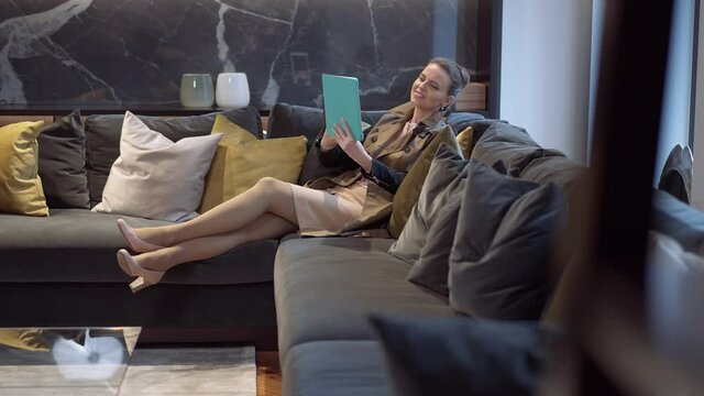 Side view wide shot gorgeous smiling elegant woman using tablet sitting on comfortable couch in furniture showroom. Portrait of Caucasian rich client choosing furnishings from online catalog