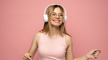 Pretty girl in a pink t-shirt and glasses listening music with her headphones and dancing on pink...