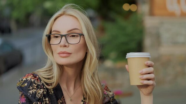 View of pretty blond woman with wide hazel eyes and eyeglasses, city center background. Business woman drinks coffee. Serious thoughtfull face. Female portrait concept