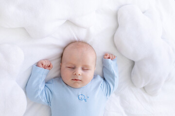 baby boy sleeps on the bed lying on his back in blue pajamas with his hands up among the pillows of clouds, healthy newborn sleep