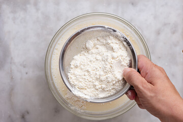 Top view of woman hand holding sieve with flour and sifting into batter in glass bowl on the marble...