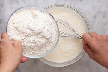 Top view of woman hand holding bowl with flour and putting into batter in glass bowl on the marble...