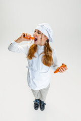 High angle view of little cute girl in white cook uniform and huge chef's hat posing isolated on...