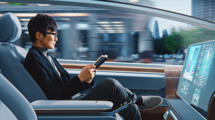 Futuristic Concept: Handsome Stylish Japanese Businessman in Glasses Reading Notebook and Watching News on Augmented Reality Screen while Sitting in a Autonomous Self-Driving Zero-Emissions Car.