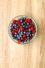 Glass bowl filled with raspberries and blueberries on wooden table. Top view.