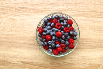 Glass bowl filled with raspberries and blueberries on wooden table. Top view.