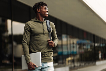 Handsome smiling dark-skinned student, wearing glasses with a backpack, holding a laptop, walking...