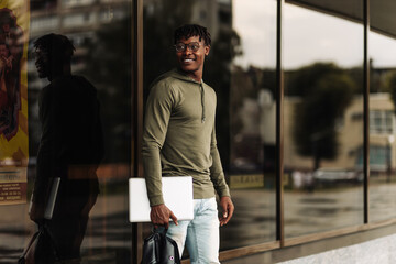 Happy smiling african american student with backpack and laptop on university background