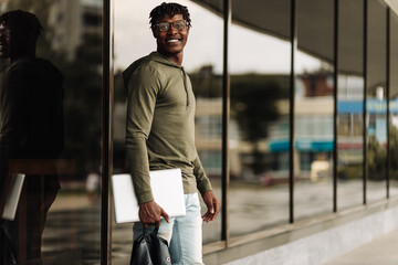 Happy smiling african american student with backpack and laptop on university background