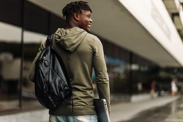 Happy smiling african american student with backpack and laptop on university background