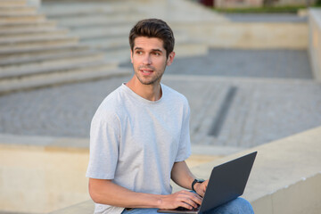 Young handsome businessman using laptop computer planning startup  looking away outdoors. Pensive...