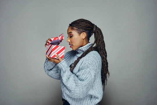 Side view of curious woman with braided black hair peeking inside Christmas gift box against gray background