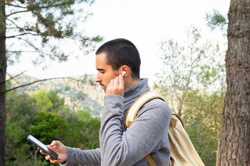 Young ethnic man using smartphone and true wireless earphones in woods