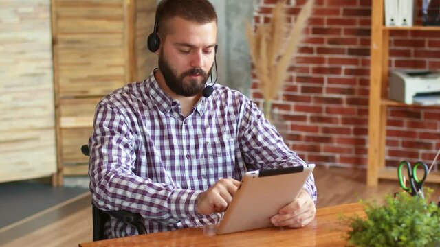 Hipster sits on wheelchair and uses electronic tablet with headset, side view. Concept of using digital gadgets and freelancing