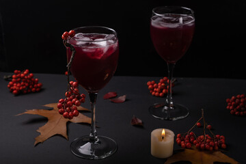 A bright red drink with ice in a glass with a beautiful rowan branch, around autumn leaves and a dying candle on a gloomy dark background. Autumn still life with halloween mood.