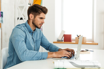 Young white man working with laptop while sitting at desk in office