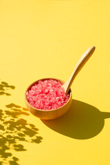 Wooden bowl with a spoon filled with pink bath sea salt. Beauty treatment for spa and wellness on yellow background. Skincare natural cosmetic concept for body care with dry plants shadows