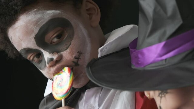 African american child in dracula decorated dresses eating lollipops and looking to his friend with black background,slow motion shot.
