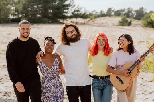 A group of very good friends are standing on the beach by the lake, of different nationalities, skin colors, holding beer bottles in their hands, playing guitar, having fun in each other's company