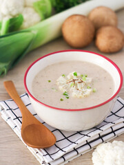 Roasted cauliflower soup in a bowl with wooden spoon and vegetables such as leek and mushroom in background. A healthy and vegan dish 