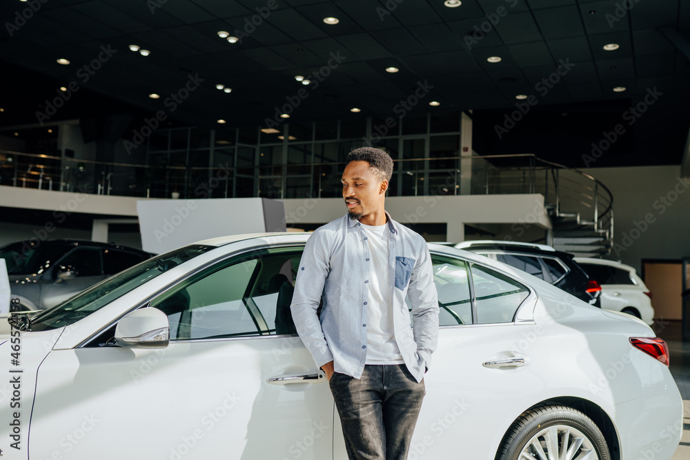 Poster Happy african man standing at dealership near white car