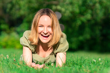 Fototapeta premium Portrait of happy cheerful positive joyful teen girl. Young beautiful woman is having fun, laughing, smiling, lying on the green grass in summer park on sunny day. Joy, happiness concept. Copy space