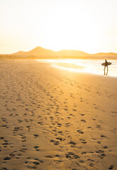 Sandy beach covered with footprints near distant surfer carrying surfboard and walking near sea at sunset © Kemedo