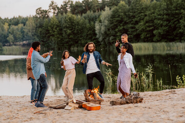 Students friends of different nationalities skin colors meet on beach by lake light bonfire fry bread sausages play guitar music dance, have fun, drink beer from bottles which they hold in their hands