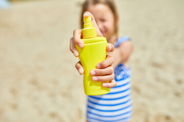 Little girl in swimsuit hold in hand yellow bottle of sunscreen standing on the beach