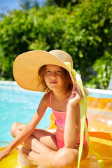 Portrait of little girl in hat relaxing in swimming pool, swims on inflatable yellow mattress