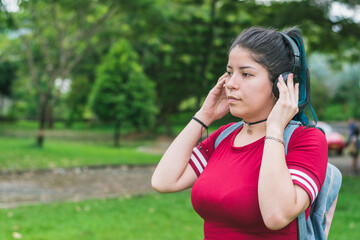 beautiful college woman with blue hair and red dress, listening to music with her wireless headphones in a park, pensive with her hands gripping the headphones. concept of freedom